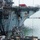 A large naval vessel, likely an aircraft carrier or amphibious assault ship, is undergoing dismantling or decommissioning operations at a shipyard. A heavy crane is lifting a large weapons or sensor system component — possibly a CIWS (Close-In Weapon System) or radar assembly — away from the ship's bow area. Multiple workers in hard hats and safety gear are supervising the lift operation from a barge or pier platform below.
