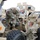 Two U.S. Army soldiers in OCP (Operational Camouflage Pattern) uniforms and blue face masks are conducting maintenance or inspection on a large military vehicle, likely an MRAP (Mine-Resistant Ambush Protected) or similar tactical wheeled vehicle. One soldier is elevated on the vehicle performing work on mechanical components, while the second soldier stands on the ground near a large off-road tire. The setting appears to be a military motor pool or staging area on a paved surface.
