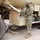 A U.S. Army soldier in OCP (Operational Camouflage Pattern) uniform and black face mask is performing maintenance or servicing on a large tan military vehicle, likely a fuel or water tanker truck. The soldier appears to be working with a hose or cable connected to a compartment on the side of the vehicle. The setting appears to be an outdoor military installation or motor pool area.