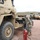 A U.S. Army soldier in OCP camouflage uniform and face mask is conducting a maintenance inspection on a tan military vehicle, likely an MRAP or heavy tactical truck. A red portable equipment unit numbered '247' — possibly a battery charger or hydraulic unit — is positioned nearby on wheeled casters. Multiple additional military vehicles are visible in the background on a gravel and dirt surface.