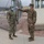 Two U.S. Army soldiers in OCP (Operational Camouflage Pattern) uniforms are shown at what appears to be a military installation checkpoint. One soldier is using a non-contact infrared thermometer to check the temperature of the other soldier. Both are wearing blue surgical face masks, consistent with COVID-19 health screening protocols.