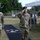 Three U.S. Army soldiers in the foreground are rendering a salute during what appears to be a military ceremony. They are wearing OCP (Operational Camouflage Pattern) uniforms, black berets, and face masks. A black runner on the ground displays 'USA NATO BDE' and 'Steadily NATO Strong' text with unit patches.