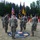 A U.S. Army military ceremony, likely a change of command or activation/inactivation ceremony, is taking place outdoors on a grass field. Soldiers in OCP (Operational Camouflage Pattern) uniforms are participating, with a color guard holding multiple flags including the U.S. flag, a unit guidon, and what appears to be a German flag. A ceremonial rug on the ground references a 'Brigade' unit with the motto 'NATO Strong.'