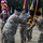 A U.S. Army change of command or activation ceremony is taking place outdoors on a grass field. Two soldiers in the foreground are exchanging a unit guidon/colors, which is the central ritual of such ceremonies. Multiple color guard soldiers stand in the background holding various national and unit flags.