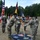 A U.S. Army change of command ceremony is taking place outdoors on a grassy field. Two senior soldiers are exchanging a unit guidon (flag) over a ceremonial rug bearing the text 'USANATO Brigade' and 'Steady! NATO'. All personnel are wearing face masks, suggesting this occurred during the COVID-19 pandemic period.