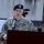 A U.S. Army Colonel is speaking at a podium equipped with a microphone. The officer wears an OCP (Operational Camouflage Pattern) uniform with a black beret bearing a blue unit flash, and a name tape reading 'GENGL' (partially visible). A video camera is visible in the background, suggesting an official military ceremony or press event.