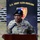 A U.S. Army soldier in camouflage uniform and black beret is speaking at a podium with a microphone. The soldier is positioned in front of a banner reading 'U.S. Army NATO Brigade' featuring a shield emblem. The setting appears to be an outdoor military ceremony or official event.
