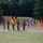 A military ceremony is taking place on a grass parade field, featuring soldiers in uniform holding or standing near multiple national and organizational flags. The display includes the American flag, German flag, and what appears to be a NATO flag among numerous other national and unit flags arranged in a row. The setting appears to be a military installation surrounded by trees, consistent with a formal change of command or allied nations ceremony.