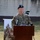 A U.S. Army senior non-commissioned officer named Valenzuela is speaking at a podium during an outdoor military ceremony. The podium bears a shield insignia consistent with U.S. Army Europe (USAREUR), and the soldier is wearing an OCP (Operational Camouflage Pattern) uniform with a black beret. A recruitment-style banner reading 'Ready to Fight' is visible in the background along with a video camera on a tripod.