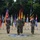 A formal military ceremony is taking place outdoors on a grass field, featuring soldiers in camouflage uniforms standing at attention with multiple national flags displayed in the background. The ceremony appears to be a NATO-related event, evidenced by a NATO logo visible on the ground mat and a NATO flag among the displayed flags. The US flag, German flag, and numerous other national flags representing NATO member nations are prominently arranged in a ceremonial arc.