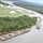 An aerial photograph showing a river bend with multiple large barges loaded with military vehicles and equipment. The barges are positioned along a wooded riverbank adjacent to what appears to be a military or industrial installation with large storage tanks, warehouses, and well-maintained grounds. The scene suggests active military logistics or equipment transport operations via inland waterway.