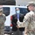 Three individuals stand outdoors near a white Ford van, with two wearing U.S. Army OCP (Operational Camouflage Pattern) uniforms and one wearing a civilian business suit. All three are wearing face masks, suggesting this photo was taken during the COVID-19 pandemic period. The uniformed personnel appear to be briefing or conversing with the civilian official.