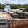Military vehicles are loaded onto a flat-deck barge being pushed by a towboat on a river. The cargo includes multiple HMMWVs (Humvees) in both tan and olive drab configurations, along with larger military trucks and covered equipment. This appears to be a military logistics or transport operation moving equipment via inland waterway.