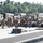 Military personnel wearing red life vests are assembled on what appears to be a pontoon bridge or floating platform near a waterway. Multiple military vehicles including HMMWVs (Humvees) and cargo trucks are staged in the background. This appears to be a military bridging or river-crossing operation or training exercise.