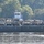 A flat barge loaded with multiple military vehicles, primarily HMMWVs (Humvees) and medium tactical trucks in tan/olive drab colors, is being pushed across a river by a civilian tugboat with a white and blue wheelhouse. Several personnel in military uniforms are visible on the barge deck among the vehicles. The scene is set on a calm river with dense green tree cover visible on the far bank.