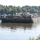 A large flat barge loaded with multiple military vehicles, including HMMWVs (Humvees) and tactical trucks in desert tan/camouflage coloring, is being pushed along a wide river by a white tugboat on the left side. A second tugboat is visible in the background on the right. The scene appears to depict a military logistics or transport operation on an inland waterway, likely in the southeastern United States based on the vegetation.