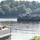 A military logistics operation is underway on a river, with a barge transporting multiple military tactical vehicles (likely HMMWVs/Humvees) across the water. A worker in a high-visibility vest and white hard hat stands on a dock or landing platform in the foreground, apparently photographing or monitoring the approaching barge. The scene suggests a river crossing or equipment transport exercise, possibly conducted by the U.S. Army.