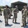 A group of military personnel in U.S. Army OCP (Operational Camouflage Pattern) uniforms are gathered outdoors on a gravel surface, appearing to be in a briefing or informal discussion. Most individuals are wearing face masks, suggesting this was taken during the COVID-19 pandemic period. The setting appears to be a military training or operational area with a metal carport structure and camouflage netting visible in the background.