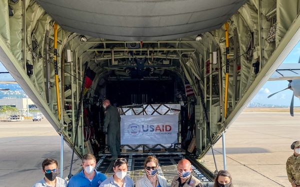 A group of six civilians wearing USAID and DART-branded clothing pose in front of an open military cargo aircraft (appears to be a C-130 Hercules) on an airport tarmac. A pallet of supplies bearing a USAID banner reading 'From the American People' is visible inside the aircraft. All individuals are wearing face masks, suggesting this was taken during the COVID-19 pandemic era.