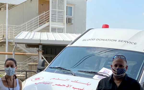 Two individuals wearing face masks stand in front of a white IVECO van marked as a 'Blood Donation Service' vehicle belonging to the Lebanese Red Cross. The vehicle displays Arabic text reading 'بنك الدم - الصليب الأحمر اللبناني' (Blood Bank - Lebanese Red Cross) along with a Red Cross emblem. The setting appears to be an urban area with buildings visible in the background.