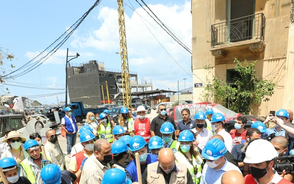 A large group of workers and officials are gathered at what appears to be a disaster relief or reconstruction site, with many wearing blue hard hats and face masks. Several individuals are holding shovels, suggesting a ceremonial groundbreaking or active debris clearing operation. USAID branding is visible on at least one hard hat, indicating international humanitarian involvement.
