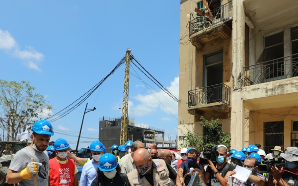 A group of workers and officials wearing blue hard hats labeled 'USAID' and 'EMT' are engaged in debris cleanup operations in what appears to be a disaster-affected urban area. The scene shows significant structural damage to surrounding buildings, with rubble and debris scattered across the street. Media personnel with cameras are present, documenting the relief and recovery efforts.