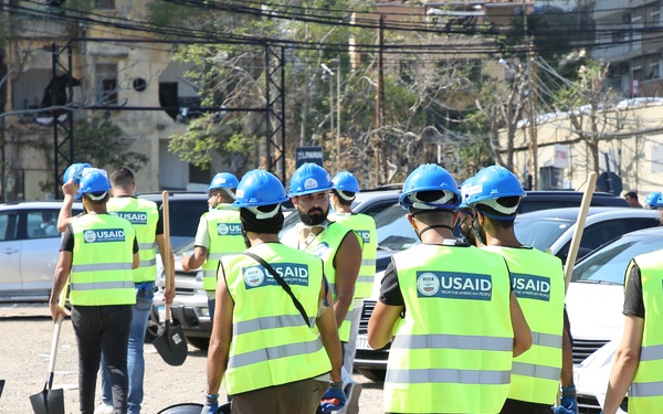 A group of USAID workers wearing bright yellow-green safety vests and blue hard hats are walking through a debris-strewn area carrying shovels, rakes, and a wheelbarrow. The scene appears to be a disaster relief or cleanup operation in an urban area with damaged surroundings and numerous parked vehicles. The workers are moving as a group, suggesting an organized humanitarian response effort.