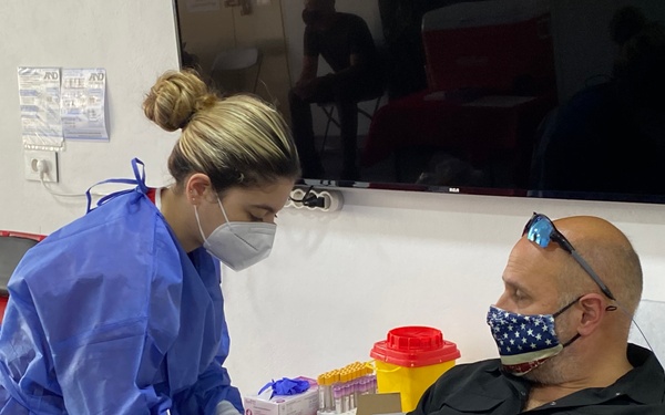 A healthcare worker in full PPE is performing a blood draw or venipuncture on a seated male patient. The setting appears to be a medical clinic or blood donation center, with both individuals wearing face masks. The scene likely takes place during or after the COVID-19 pandemic period based on the PPE usage.