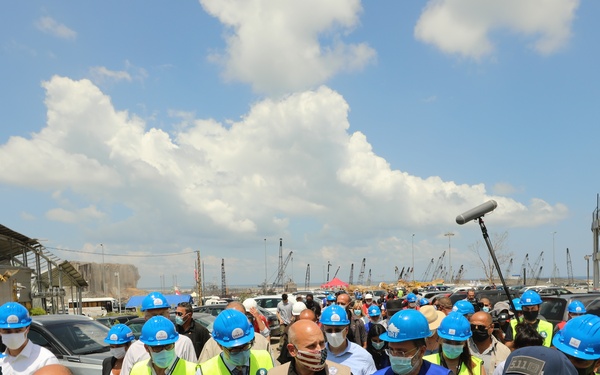 A large group of officials and aid workers wearing blue hard hats and high-visibility yellow vests are walking through a damaged port area, consistent with a post-disaster assessment or inspection visit. Several individuals wear face masks and identification badges, with at least one person wearing a USAID-branded vest. The surrounding environment shows heavily damaged structures, debris, and port cranes visible in the background.