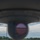 The image is taken from beneath an aircraft, looking along the fuselage toward the nose or tail section. A prominent rounded sensor dome or light fixture is visible in the center foreground, with an aircraft wing visible to the left. The background shows an airport environment with light poles, trees, and a partly cloudy sky.