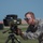 A U.S. Air Force Technical Sergeant in ABU (Airman Battle Uniform) is operating a tripod-mounted military optical/laser device outdoors on what appears to be an airfield or base. The servicemember is wearing protective eyewear and tactical gloves while closely examining or adjusting the equipment. The setting appears to be a clear day with open terrain visible in the background.