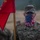 A U.S. Army soldier named Benedicks stands at attention holding a red military guidon flag. The soldier is wearing OCP (Operational Camouflage Pattern) uniform with a U.S. flag-patterned gaiter/face covering. The image appears to be taken during a military formation or ceremony in overcast, possibly rainy conditions.
