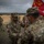 Two U.S. Army soldiers in OCP (Operational Camouflage Pattern) uniforms are engaged in what appears to be a guidon transfer ceremony on a military training range. The soldier facing the camera has the name tape 'ROSARIO' and a U.S. Army patch, while the soldier holding the red guidon has the name tape 'HARRIS'. Additional soldiers and military vehicles are visible in the background under overcast skies.