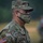 A U.S. Army soldier identified as 'Rosario' is photographed in profile view wearing Operational Camouflage Pattern (OCP) uniform and a matching camouflage face mask with their name printed on it. The soldier appears to be outdoors in rainy conditions, with a blurred second individual visible in the background. The image is a close-up portrait-style photograph taken in a field or open outdoor environment.