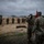 A military formation ceremony is taking place on an open sandy training ground under overcast skies. Two soldiers in the foreground are saluting toward a formation of troops standing at attention, with one soldier holding a red unit guidon flag. Smoke or dust is visible in the background near military vehicles, suggesting recent artillery or training activity.