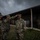A group of U.S. Army soldiers in OCP (Operational Camouflage Pattern) uniforms are gathered outdoors near a large military building under overcast skies. A soldier in the foreground is rendering a salute, suggesting a formal military ceremony or inspection is taking place. All personnel are wearing face masks, indicating this event occurred during the COVID-19 pandemic period.