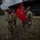 A group of U.S. Army soldiers in OCP (Operational Camouflage Pattern) uniforms are participating in what appears to be a military ceremony, likely a change of command or guidon transfer, at an outdoor training facility. Several soldiers are wearing face masks, suggesting the event took place during the COVID-19 pandemic period. A red unit guidon flag with yellow insignia is prominently displayed during the ceremony.
