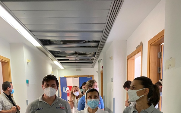 Two USAID DART (Disaster Assistance Response Team) personnel in gray polo shirts are walking through a hospital corridor alongside a medical professional in a white coat. All individuals are wearing face masks, suggesting a health-conscious or pandemic-era context. The corridor shows signs of structural damage, with a partially collapsed ceiling visible in the background.
