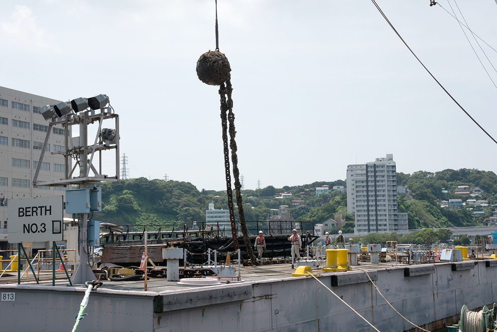 A harbor scene showing workers on a dock or vessel at Berth No. 3, with a crane lifting what appears to be a heavily encrusted anchor or large marine object covered in marine growth. The setting appears to be a Japanese port facility, with residential and commercial buildings visible on a hillside in the background. Several workers in hard hats and safety gear are visible on the deck overseeing the operation.