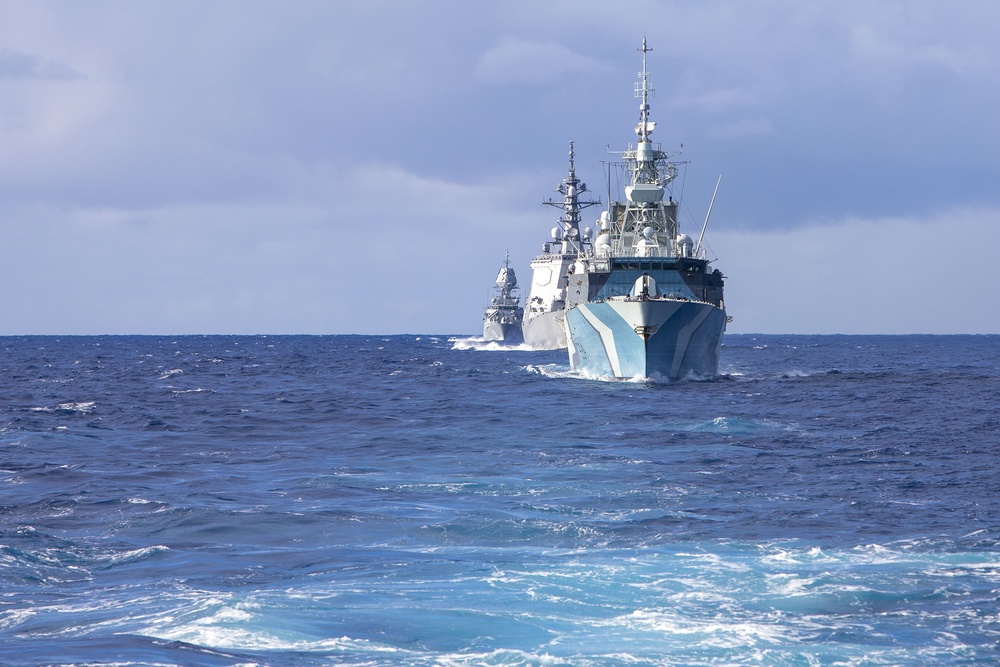 The image shows a formation of military naval vessels underway on open ocean waters. At least three warships are visible sailing in a column formation, with the closest vessel displaying a distinctive blue and white geometric camouflage pattern on its bow. The sky is partly cloudy with blue tones, and the sea shows moderate wave activity.