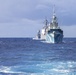 The image shows a formation of military naval vessels underway on open ocean waters. At least three warships are visible sailing in a column formation, with the closest vessel displaying a distinctive blue and white geometric camouflage pattern on its bow. The sky is partly cloudy with blue tones, and the sea shows moderate wave activity.