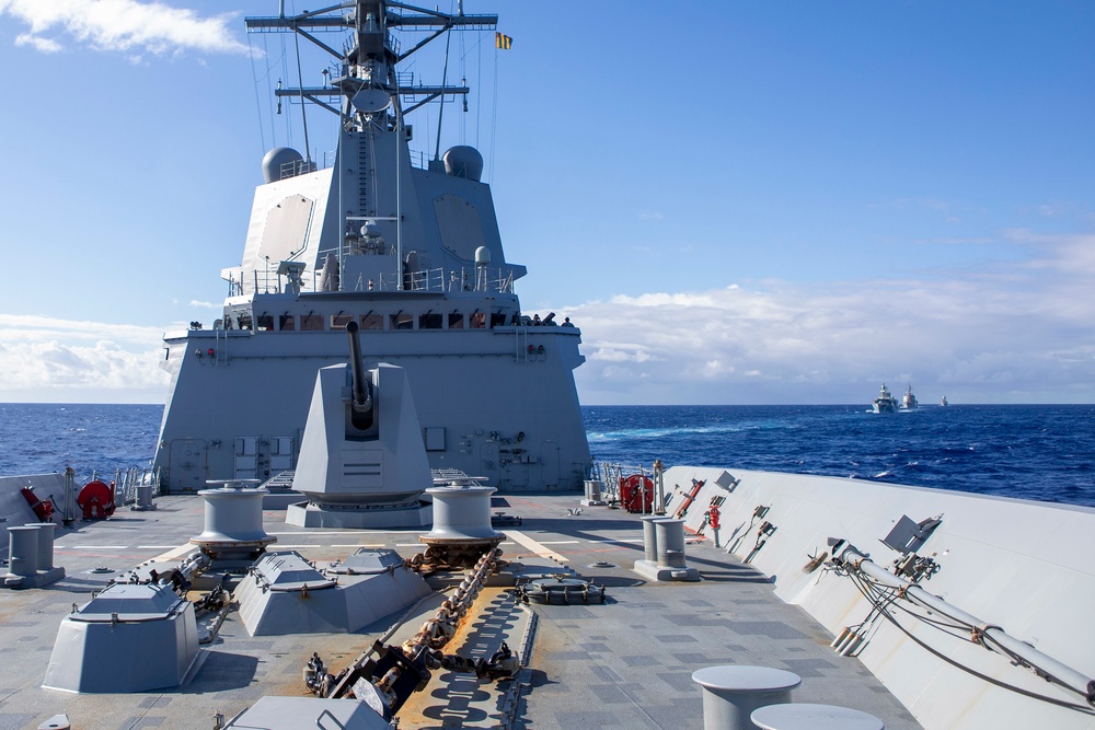 The image shows the forward deck of a modern naval warship underway at sea, featuring a prominent deck gun and superstructure with radar domes and mast equipment. The vessel appears to be part of a formation, with at least two additional ships visible on the horizon in the background. The ship displays a grey naval paint scheme consistent with contemporary Western naval vessels.
