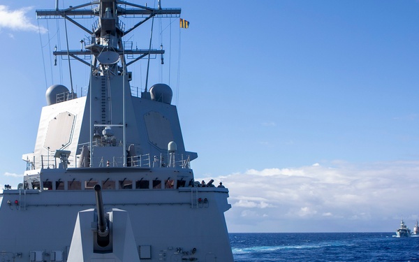 The image shows the forward deck of a modern naval warship underway at sea, featuring a prominent deck gun and superstructure with radar domes and mast equipment. The vessel appears to be part of a formation, with at least two additional ships visible on the horizon in the background. The ship displays a grey naval paint scheme consistent with contemporary Western naval vessels.
