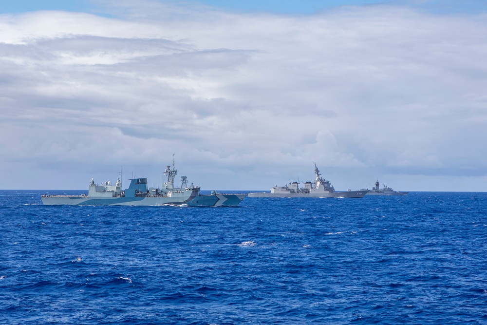 The image shows a formation of four naval warships sailing together on open blue ocean waters under a partly cloudy sky. The vessels appear to be from different navies operating in a joint or multinational exercise formation. Hull numbers are partially visible on at least two of the ships, including what appears to be '334' and '174'.