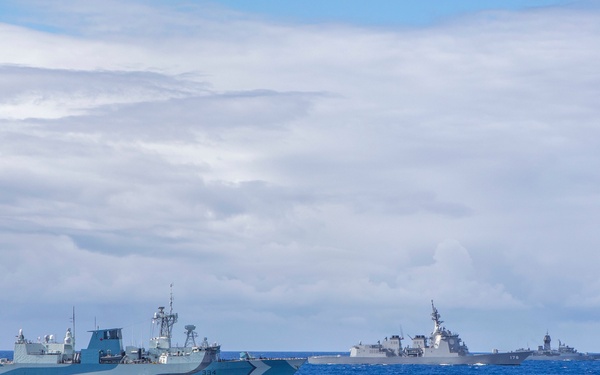 The image shows a formation of four naval warships sailing together on open blue ocean waters under a partly cloudy sky. The vessels appear to be from different navies operating in a joint or multinational exercise formation. Hull numbers are partially visible on at least two of the ships, including what appears to be '334' and '174'.