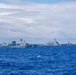 The image shows a formation of four naval warships sailing together on open blue ocean waters under a partly cloudy sky. The vessels appear to be from different navies operating in a joint or multinational exercise formation. Hull numbers are partially visible on at least two of the ships, including what appears to be '334' and '174'.