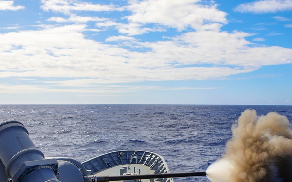 A naval warship is actively firing its deck-mounted gun cannon over open ocean waters. A large cloud of gray-brown smoke and muzzle blast is visible at the gun barrel's muzzle, indicating a live fire exercise or combat operation. The image captures the moment of discharge with the projectile already having left the barrel.