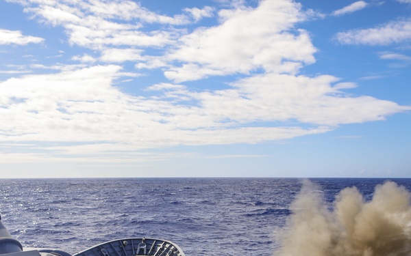 A naval warship is captured mid-firing of its main deck gun, with a large cloud of propellant smoke and a visible projectile streak extending from the muzzle. The photograph is taken from an elevated position on the ship's superstructure, looking forward along the deck toward the bow. The vessel is underway on open blue ocean under partly cloudy skies.