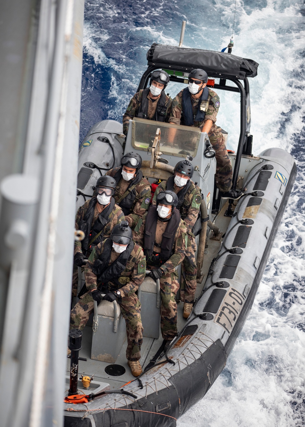 A rigid inflatable boat (RIB) carrying approximately six to seven military personnel in camouflage uniforms approaches what appears to be a larger vessel. All personnel are wearing black helmets, protective goggles, face masks, and life vests, suggesting a maritime boarding or training operation. The boat bears a visible hull marking 'F731-01' and insignia consistent with a naval force.
