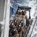 A rigid inflatable boat (RIB) carrying approximately six to seven military personnel in camouflage uniforms approaches what appears to be a larger vessel. All personnel are wearing black helmets, protective goggles, face masks, and life vests, suggesting a maritime boarding or training operation. The boat bears a visible hull marking 'F731-01' and insignia consistent with a naval force.